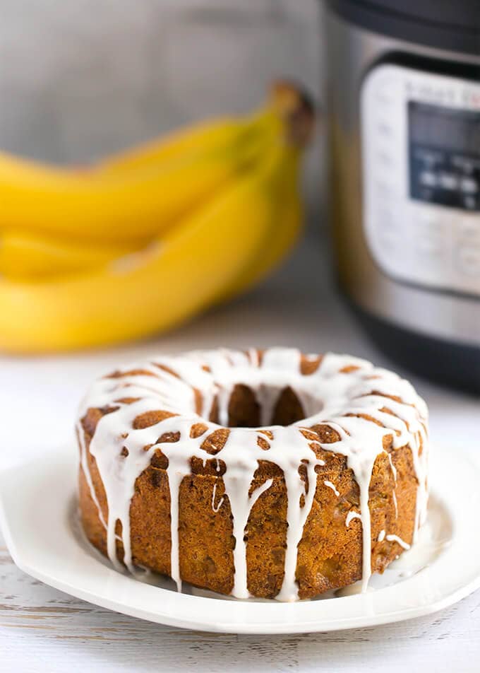 Bunt loaf of Banana Bread with vanilla icing glaze, all on a white plate in front of a pressure cooker