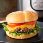 Hamburgers with cheese, lettuce, and tomato on a black plate in front of a pressure cooker