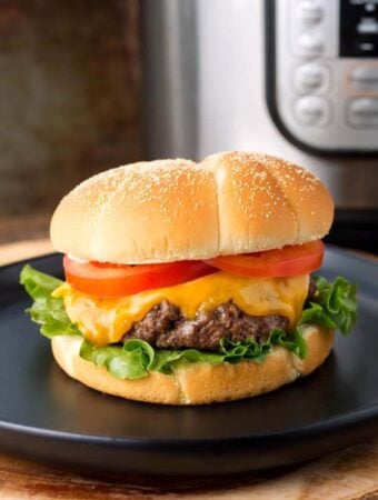 Hamburgers with cheese, lettuce, and tomato on a black plate in front of a pressure cooker