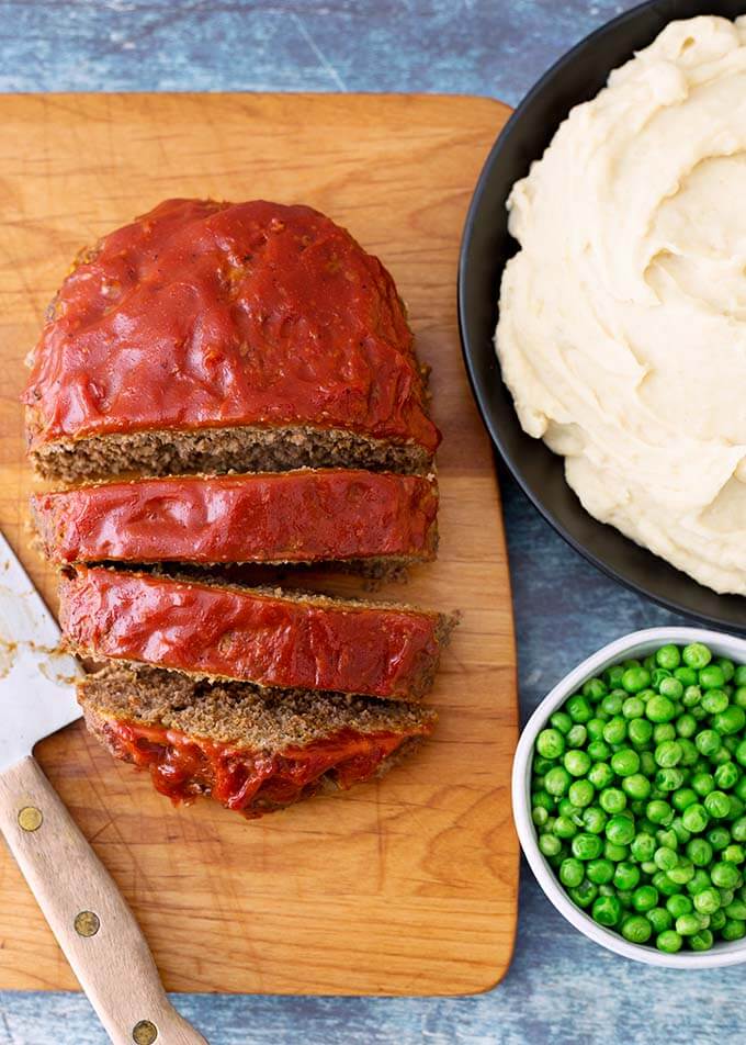 Sliced Meatloaf on a wooden cutting board next to a black bowl of mashed potatoes and a white bowl of peas
