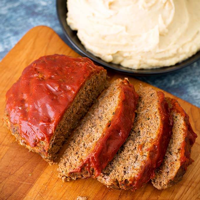 Sliced Meatloaf on a wooden cutting board with a black bowl full Mashed Potatoes