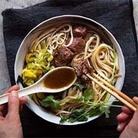 Taiwanese Beef Noodle Soup in a white bowl with fingers holding white soup spoon and chopsticks