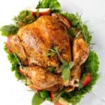 Top view of a cooked Whole Chicken on a white plate with green herbs, leafy vegetables, and apple slices