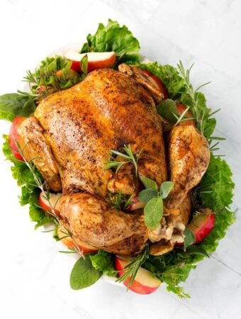 Top view of a cooked Whole Chicken on a white plate with green herbs, leafy vegetables, and apple slices