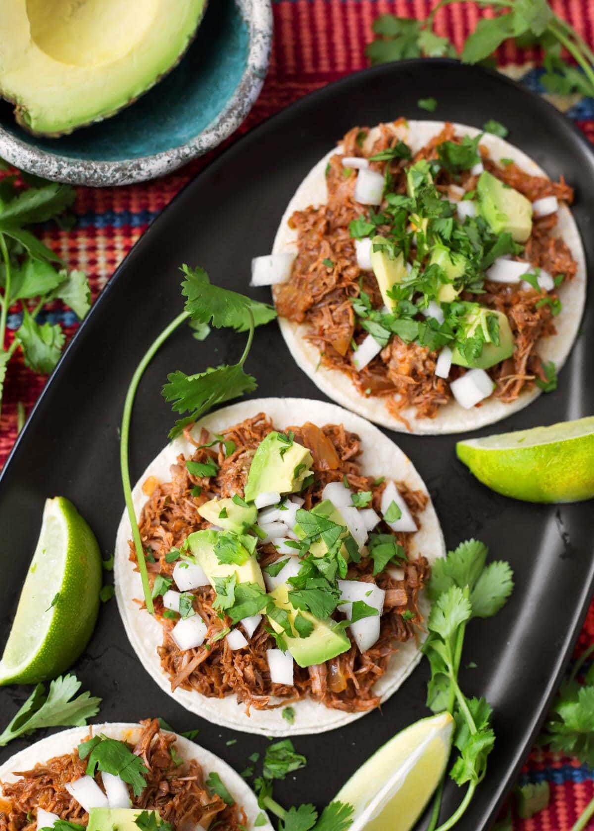 Barbacoa Beef on white corn tortillas topped with chopped avocados, onion and cilantro on a black oblong platter