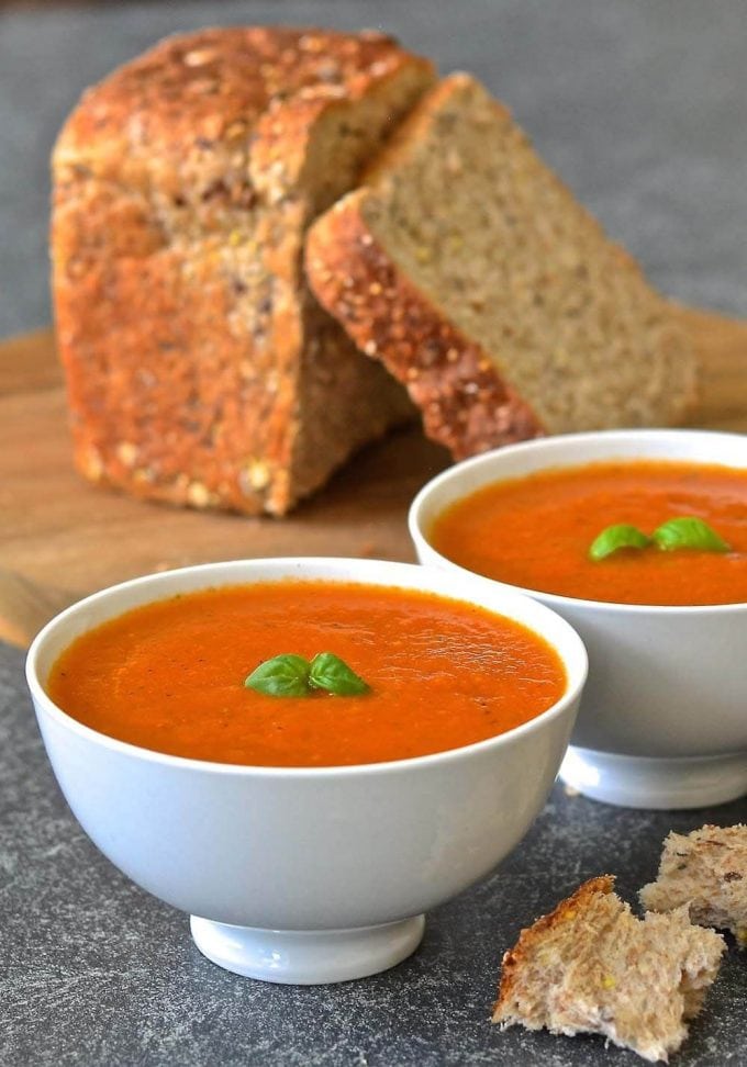 Two bowls of easy tomato basil soup in front of a loaf of bread on a wooden cutting board