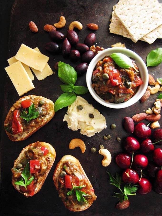 Ratatouille in a small white bowl with some spread on french bread on a platter with different cheeses, olives, cherries, and nuts