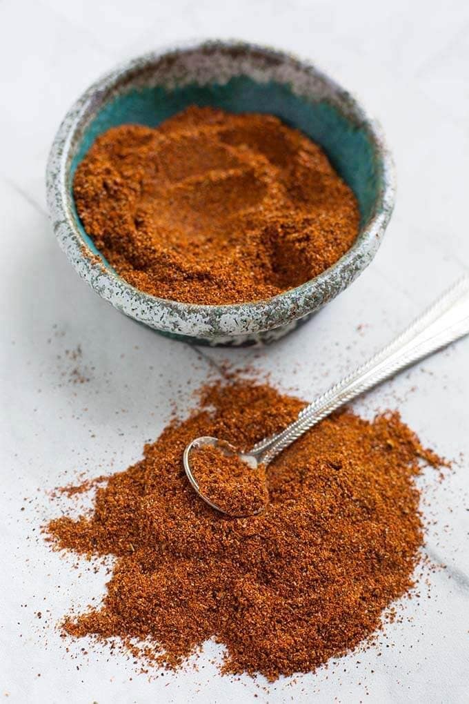 Homemade Fajita Seasoning in a turquoise stoneware bowl next to a small pile of seasoning with a silver teaspoon on a white background