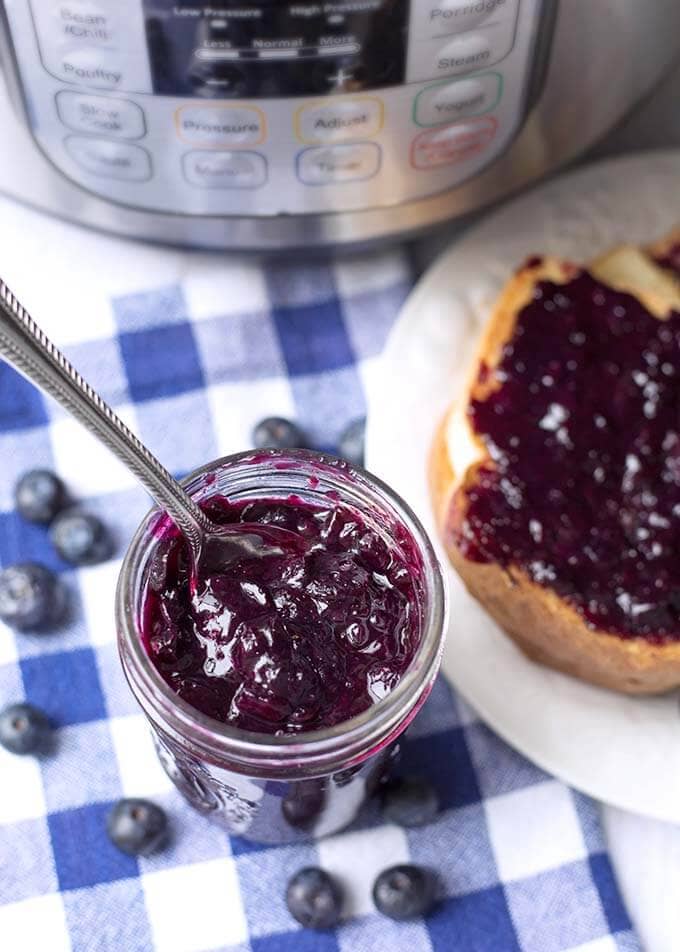 Blueberry Jam in a small glass jar with a silver spoon, next to a white plate with jam on bread