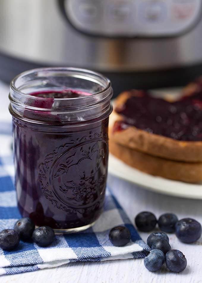 Jar of blueberry jam in front of bread with jam and pressure cooker