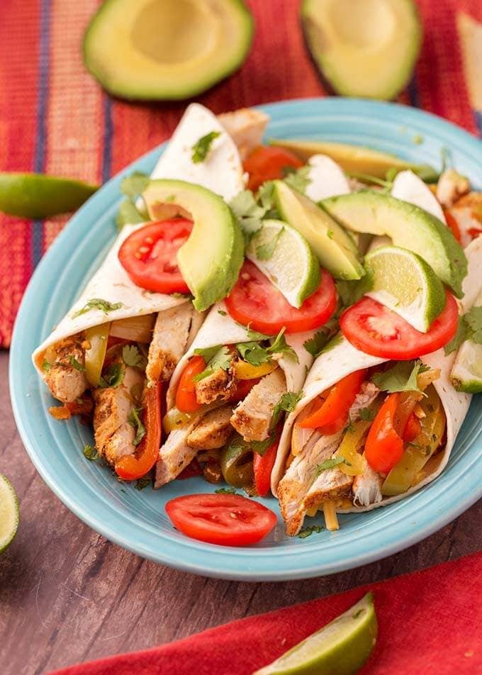 Three Chicken Fajitas on a blue plate topped with sliced avocados, tomatoes, and limes