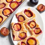 Four slices of Fresh Plum Cake, also called Pflaumenkuchen and Zwetschgenkuchen (German Plum Cake) on a white oblong plate next to baking sheet with remaining dish