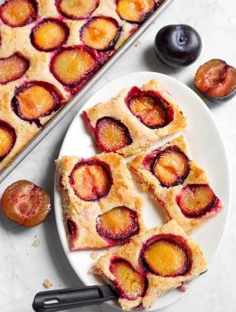 Four slices of Fresh Plum Cake, also called Pflaumenkuchen and Zwetschgenkuchen (German Plum Cake) on a white oblong plate next to baking sheet with remaining dish