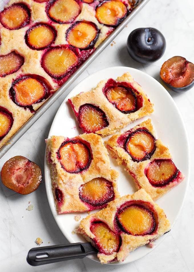 Four slices of Fresh Plum Cake, also called Pflaumenkuchen on a white oblong plate, next to a baking sheet of remaining dish