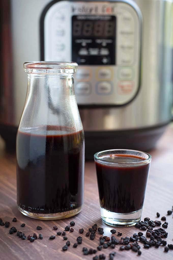 Small bottle of Elderberry Syrup next to a small glass next to dried elderberries in front of a pressure cooker