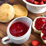 Strawberry Sauce in a small white pitcher next to biscuits with strawberry syrup spread on them next to a white colander full of halved strawberries