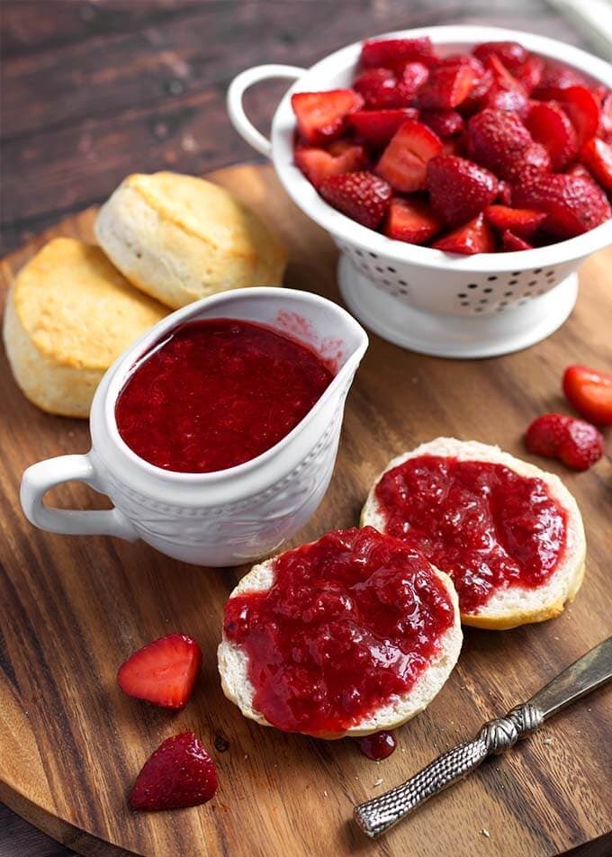 Strawberry Sauce in a small white pitcher next to biscuits with strawberry syrup spread on them next to a white colander full of halved strawberries