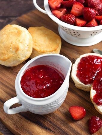 Strawberry Sauce in a small white pitcher next to biscuits with strawberry syrup spread on them next to a white colander full of halved strawberries
