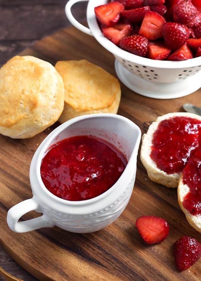 Strawberry Sauce in a small white pitcher next to biscuits with strawberry syrup spread on them next to a white colander full of halved strawberries