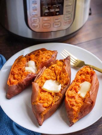 Three cooked sweet potatoes with butter and a fork on a white plate in front of a pressure cooker