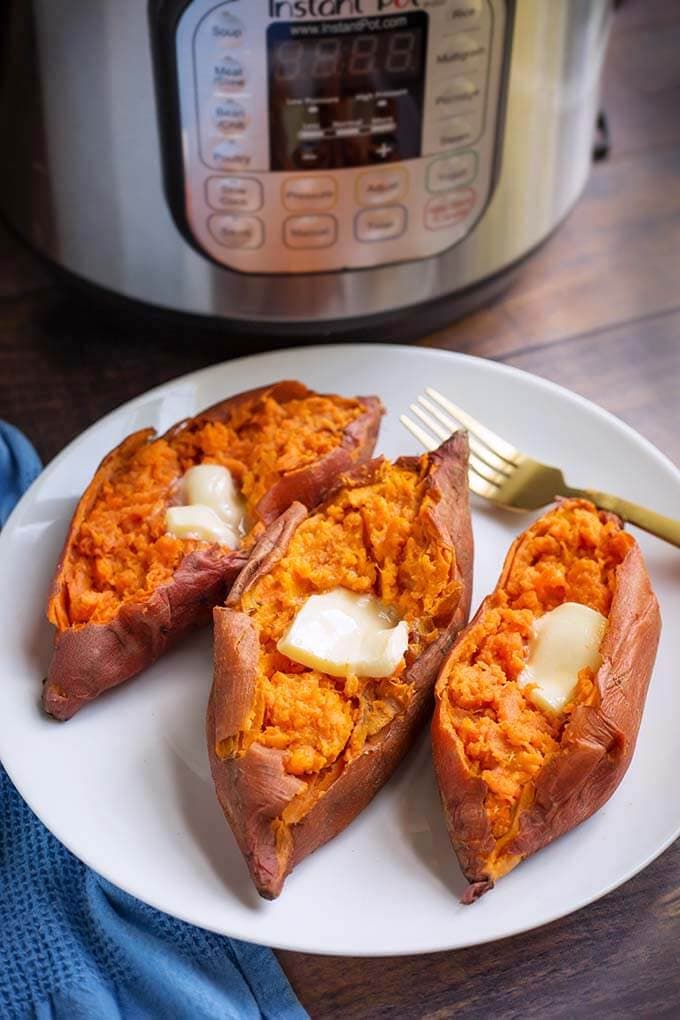 Three cooked sweet potatoes with butter and a fork on a white plate in front of a pressure cooker