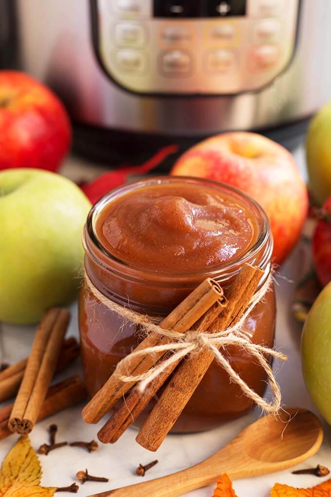 Small glass jar of Apple Butter with cinnamon sticks tied with twine in front of green and red apples and a pressure cooker