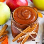 Small glass jar of Apple Butter with cinnamon sticks tied with twine in front of green and red apples and more cinnamon sticks
