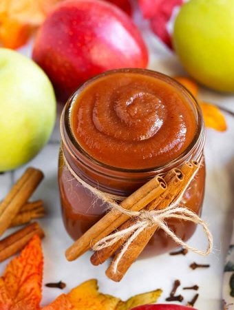 Small glass jar of Apple Butter with cinnamon sticks tied with twine in front of green and red apples and more cinnamon sticks