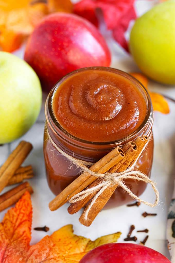 Small glass jar of Apple Butter with cinnamon sticks tied with twine in front of green and red apples and more cinnamon sticks