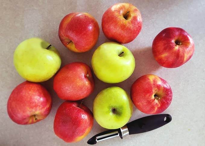 Several red and green apples next to a vegetable peeler