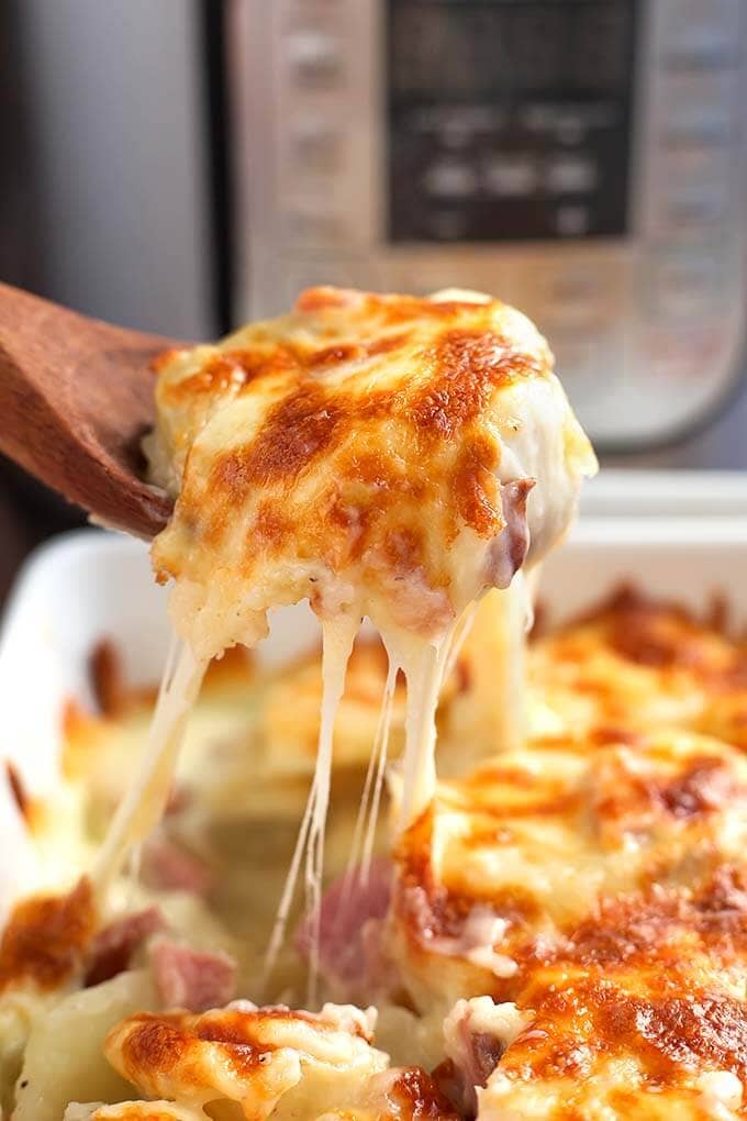 Closeup of wooden spoon scooping up Scalloped Potatoes in a square white baking dish in front of a pressure cooker