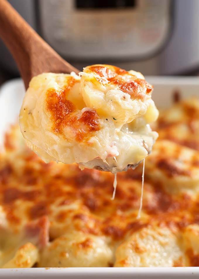 Closeup of a wooden spoon scooping scalloped potatoes from white baking dish in front of a pressure cooker