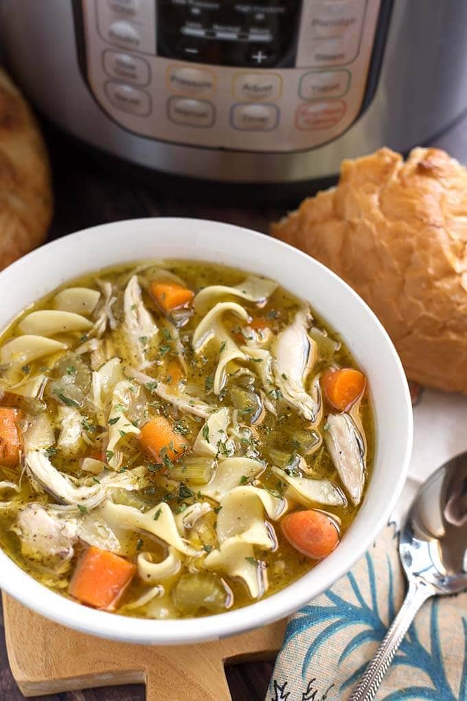 Top view of Chicken Noodle Soup in a white bowl on top of a wooden board in front of a pressure cooker