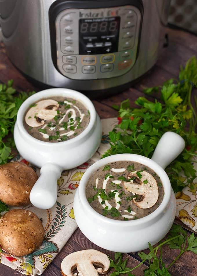 Two white handled bowls of Mushroom Soup next to parsley and mushrooms in front of a pressure cooker