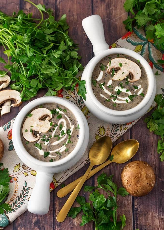 Top view of two white handled bowls of Mushroom Soup recipe next to two golden spoons,parsley and mushrooms