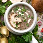 Top view of Mushroom Soup in a white handled bowl topped with a fresh mushroom slice and parsley