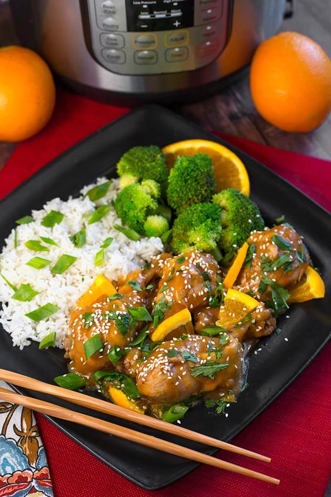 Orange Chicken Legs with rice, broccoli, and an orange slice on a square black plate with chopsticks, in front of a pressure cooker