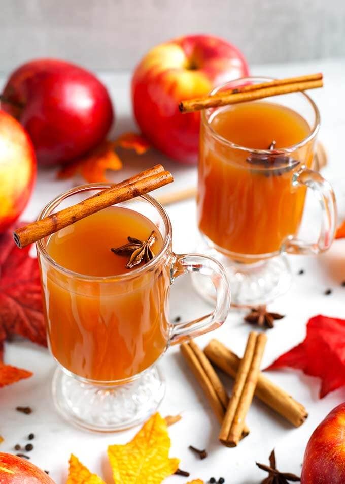 Two Spiced Apple Hot Toddy in glasses topped with cinnamon sticks and anise seeds, surrounded by cinnamon sticks, whole cloves and anise seeds as well as apples and fall leaves