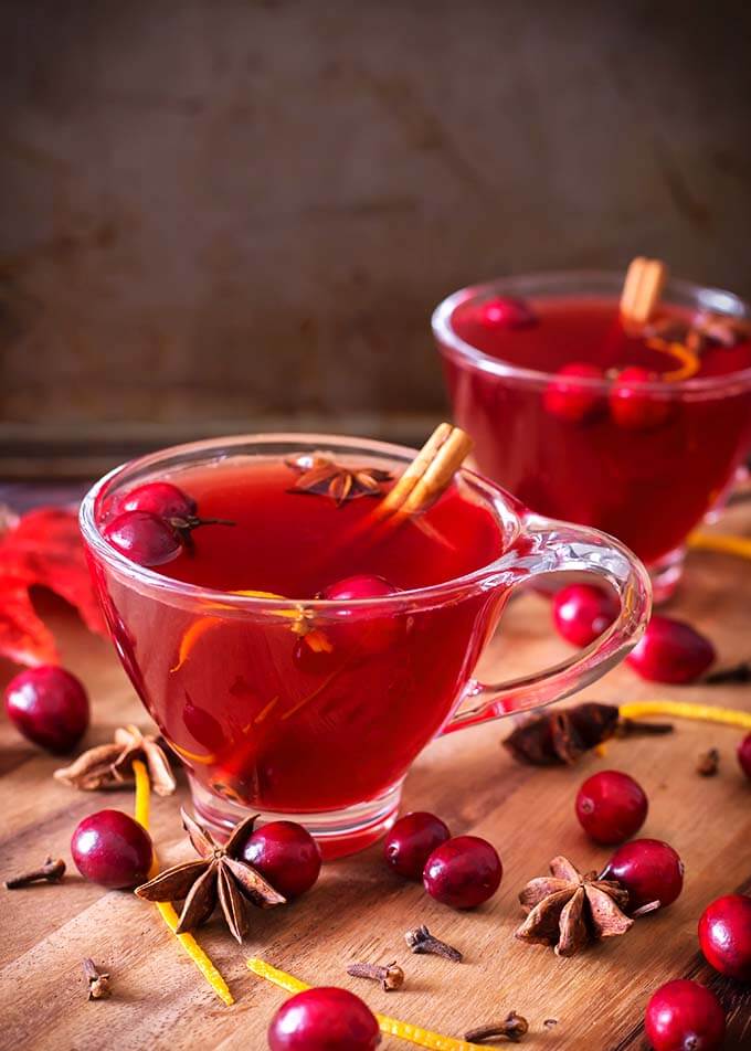 Two Spiced Cranberry Hot Toddy with whole cranberries, cinnamon stick, orange zest, and anise in glasses on a wooden board