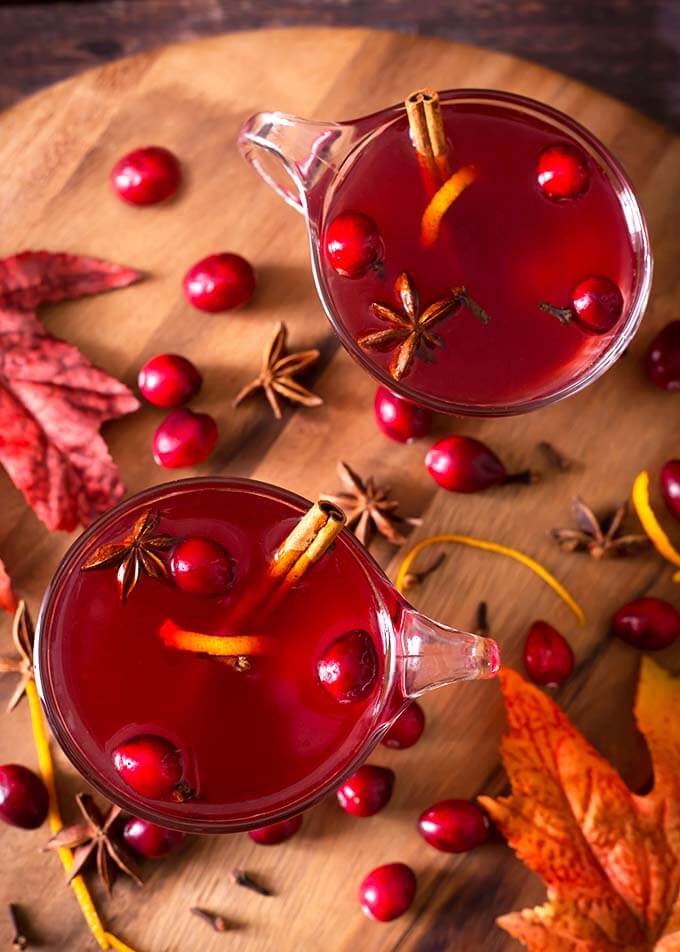 Top view of two Spiced Cranberry Hot Toddy with cinnamon sticks, whole cranberries, and anise seed in glasses on a round wooden board