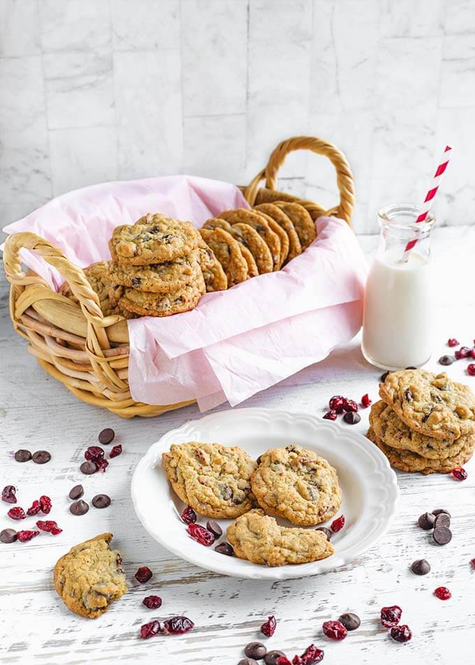 Three Cranberry Dark Chocolate Cookies on a white plate in front of a oblong basket lined with a pink napkin with several cookies inside as well as a glass of milk with a red and white stripped straw with other cookies stacked all on a white wood background