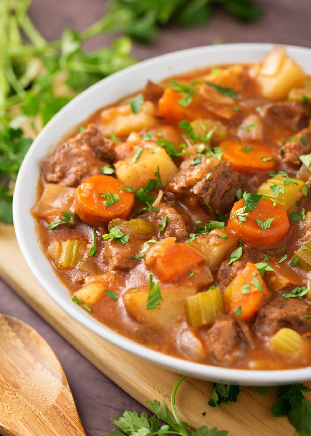 Beef Stew in a white bowl on a wooden board next to fresh parsley