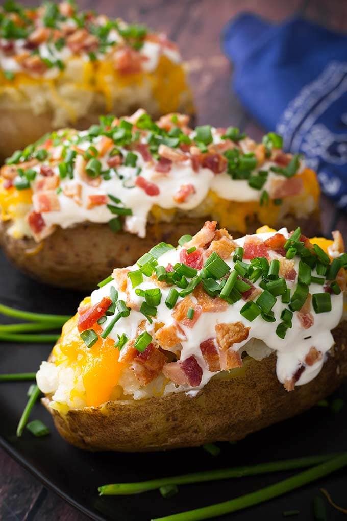 Three Baked Potato topped with cheese, bacon, sour cream and chives on wooden board with green onions