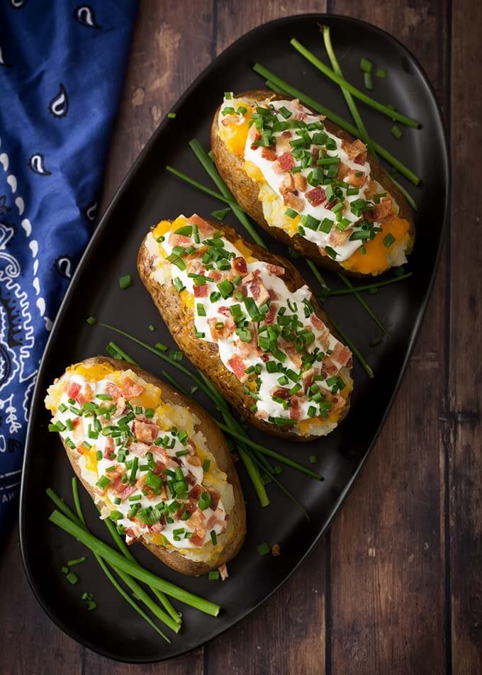 Top view of three Baked Potatoes topped with cheese, sour cream, chopped green oinion, and bacon bits on a oblong black platter