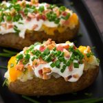 Two Baked Potatoes topped with sour cream, cheese, bacon bits and chopped green onion on a black plate