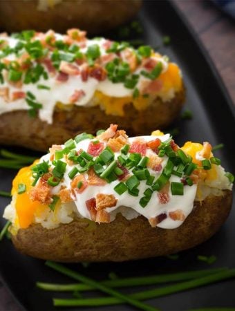 Two Baked Potatoes topped with sour cream, cheese, bacon bits and chopped green onion on a black plate