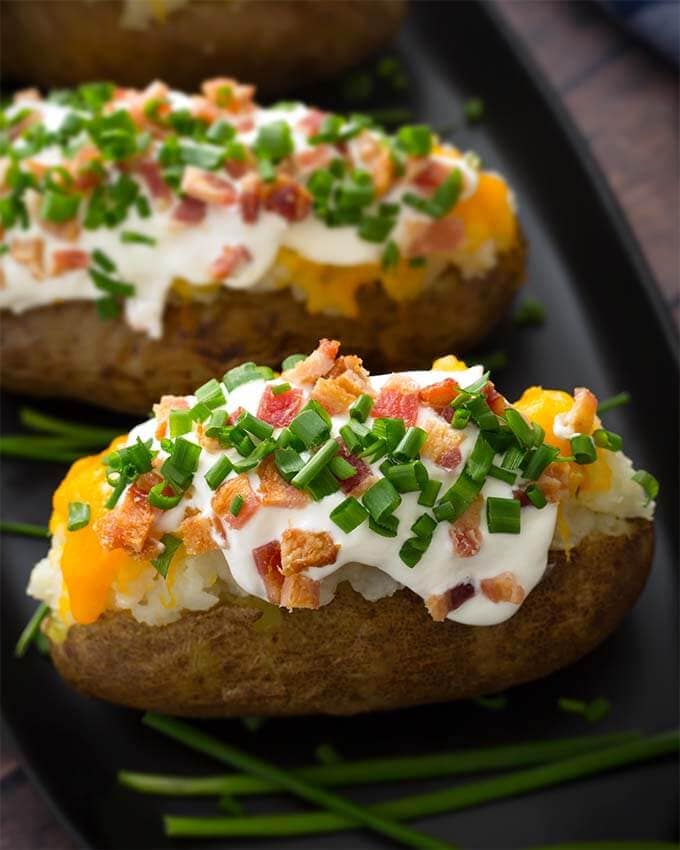 Two Baked Potatoes topped with sour cream, cheese, bacon bits and chopped green onion on a black plate