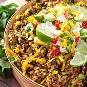 Beef Taco Bowls in a beige bowl topped with sliced lime, shrdded cheese, diced avocado and tomato and shredded cheese