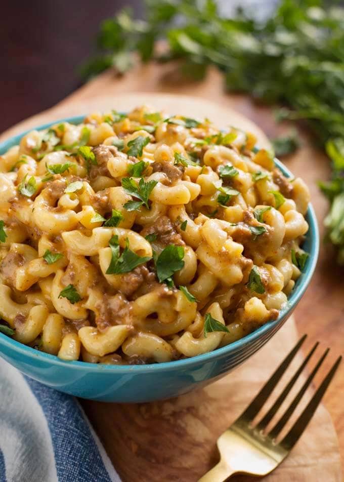 Cheeseburger Macaroni in a blue bowl next to a gold fork all on a wooden board