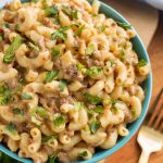 Cheeseburger Macaroni in a blue bowl next to a gold fork all on a wooden board